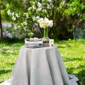 May include: A white linen tablecloth with a bouquet of white and yellow daffodils in a glass vase on a table with a stack of white plates and a place card with the number 8. The table is set in a garden with green grass and white flowers.