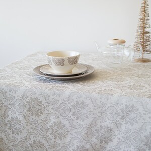 May include: A white tablecloth with a gray damask pattern. A white bowl and two white plates are on the tablecloth. A glass teapot and a small decorative tree are on the table.