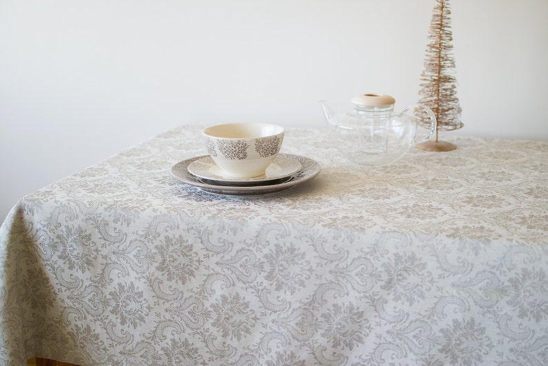 May include: A white tablecloth with a gray damask pattern. A white bowl and two white plates are on the tablecloth. A glass teapot and a small decorative tree are on the table.