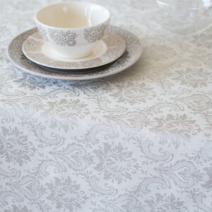 May include: A white tablecloth with a gray damask pattern. A white teacup and saucer are on top of a stack of two plates. The teacup has a gray floral design.