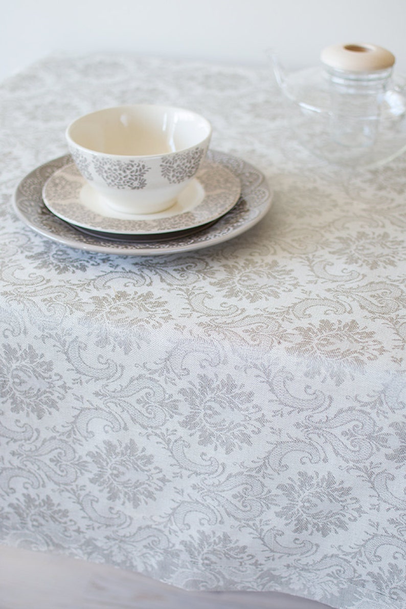 May include: A white tablecloth with a gray damask pattern. A white teacup and saucer are on top of a stack of two plates. The teacup has a gray floral design.