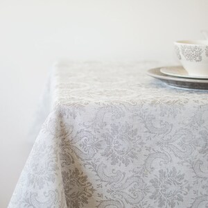 May include: A white tablecloth with a gray floral pattern. The tablecloth is draped over a table and has a slightly textured surface.