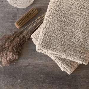 Puede incluir: Dos toallas beige con tejido de gofre apiladas sobre una superficie de madera envejecida. Un sombrero pequeño y claro, un cepillo de madera y hierba ornamental seca se disponen cerca, creando una estética rústica y de spa.
