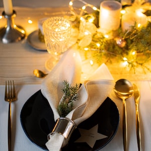 Può includere: Elegante mise en place con piatto nero, posate dorate e tovagliolo di lino. Un rametto di verde è inserito nel portatovagliolo. Candele e luci di fata creano un'atmosfera calda.