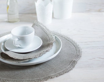 May include: A white ceramic teacup and saucer on a gray linen placemat with a lace trim. The placemat is on a white wooden table.