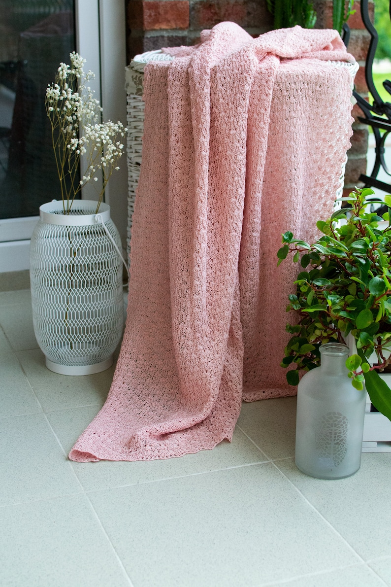 K&ouml;nnte beinhalten: Eine rosa geh&auml;kelte Wolldecke, die &uuml;ber einen wei&szlig;en Korb aus Weidengeflecht gelegt ist. Die Decke hat ein zartes, offenes Webmuster. Der Korb steht auf einem gefliesten Boden.