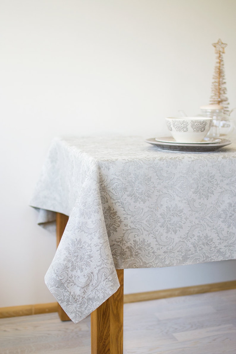 May include: A white linen tablecloth with a gray floral pattern, draped over a wooden table. A white teacup and saucer are on the table, along with a small decorative tree.