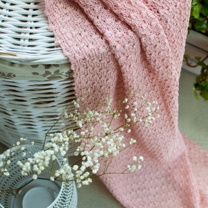 K&ouml;nnte beinhalten: Eine rosa gestrickte Decke, die &uuml;ber einen wei&szlig;en Weidenkorb gelegt ist, mit einer wei&szlig;en Metallvase gef&uuml;llt mit wei&szlig;en Babyschnaps-Blumen.