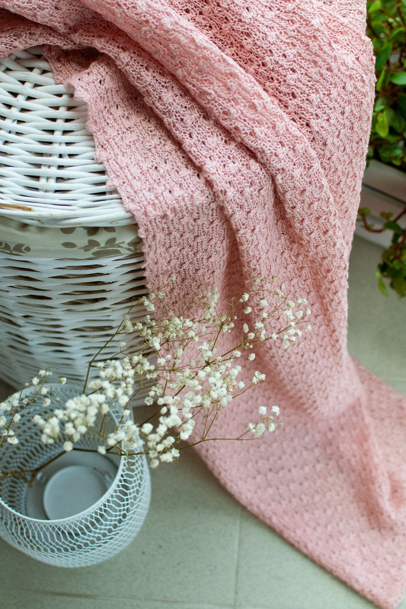 K&ouml;nnte beinhalten: Eine rosa gestrickte Decke, die &uuml;ber einen wei&szlig;en Weidenkorb gelegt ist, mit einer wei&szlig;en Metallvase gef&uuml;llt mit wei&szlig;en Babyschnaps-Blumen.