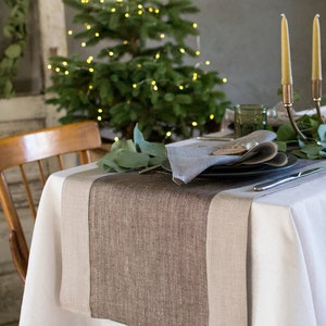 Peut inclure: Une nappe blanche avec un chemin de table en lin brun et beige. Le chemin de table a un tissage texturé. Une assiette, une serviette et des couverts sont disposés sur la table. Un petit sapin de Noël avec des lumières blanches est en arrière-plan.