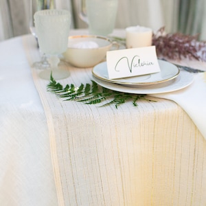 Könnte beinhalten: Ein weißer Tisch mit einem goldgestreiften Läufer. Ein weißer Teller mit goldenem Rand liegt auf dem Läufer. Eine weiße Karte mit dem Namen "Victoria" in schwarzer Schrift liegt auf dem Teller.