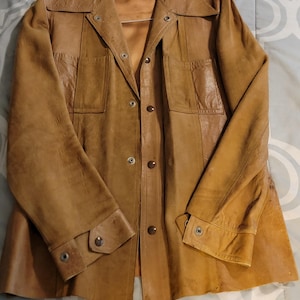 Puede incluir: Chaqueta de cuero marrón vintage con cuello, cierre de botones y dos bolsillos delanteros. La chaqueta tiene mangas largas con puños abotonados y está colgada de una percha de alambre. El cuero tiene un aspecto desgastado y texturizado.