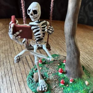 May include: A whimsical figurine of a skeleton on a swing, holding a small red device, possibly a phone or tablet. The skeleton is set against a backdrop of a miniature forest with red and white mushrooms and a tree trunk.