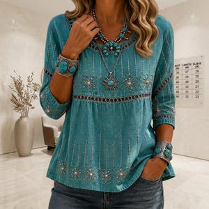 May include: A teal blue long-sleeved top featuring white embroidered floral patterns and vertical dotted lines. The garment is worn with a silver-toned necklace and two matching silver-toned cuff bracelets, each set with three oval turquoise-coloured stones.