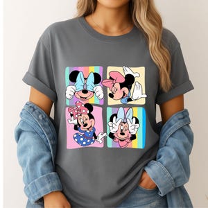Może przedstawiać: Szara koszulka z kolorową grafiką Myszki Minnie w czterech r&oacute;żnych pozach. Każdy kwadrat przedstawia Minnie z inną kokardą i wyrazem twarzy, na tle pastelowego tęczowego tła. Koszulka ma podwinięte rękawy.