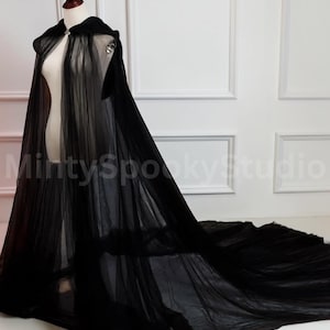 May include: A floor-length black hooded cape made of sheer, layered tulle fabric. The garment features a hood, a silver-toned metal clasp at the neckline, and a long, flowing train that extends across the floor. The text MintySpookyStudio is visible.