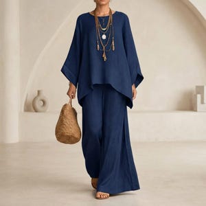 Könnte beinhalten: Ein dunkelblaues zweiteiliges Set bestehend aus einem locker sitzenden Langarmshirt und einer weiten Hose. Das Outfit wird durch eine mehrlagige goldfarbene Anhängerkette und eine strukturierte, hellbraune geflochtene Handtasche ergänzt.