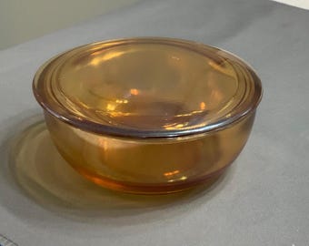 May include: A round, amber-colored glass bowl with a matching lid. The bowl has a slightly flared shape and a smooth, reflective surface. The lid fits snugly on top, creating a complete and functional piece. The glass has a warm, golden hue.