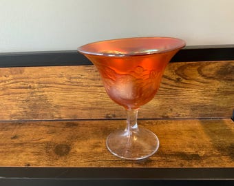 May include: An orange iridescent glass goblet featuring a wide, flared rim and a pedestal base. The exterior surface of the bowl displays a molded relief pattern of floral motifs, creating a textured appearance with a reflective, metallic sheen.