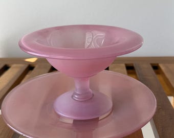 May include: A frosted pink glass compote dish featuring a wide, shallow bowl supported by a central pedestal base, which is attached to a matching circular glass plate. The glass has a smooth, matte finish with a translucent, soft pink hue.