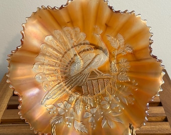 May include: An orange iridescent carnival glass bowl with a ruffled edge. The center features a molded relief design of a peacock with a fanned tail perched on a fence, surrounded by embossed floral and leaf patterns across the surface.