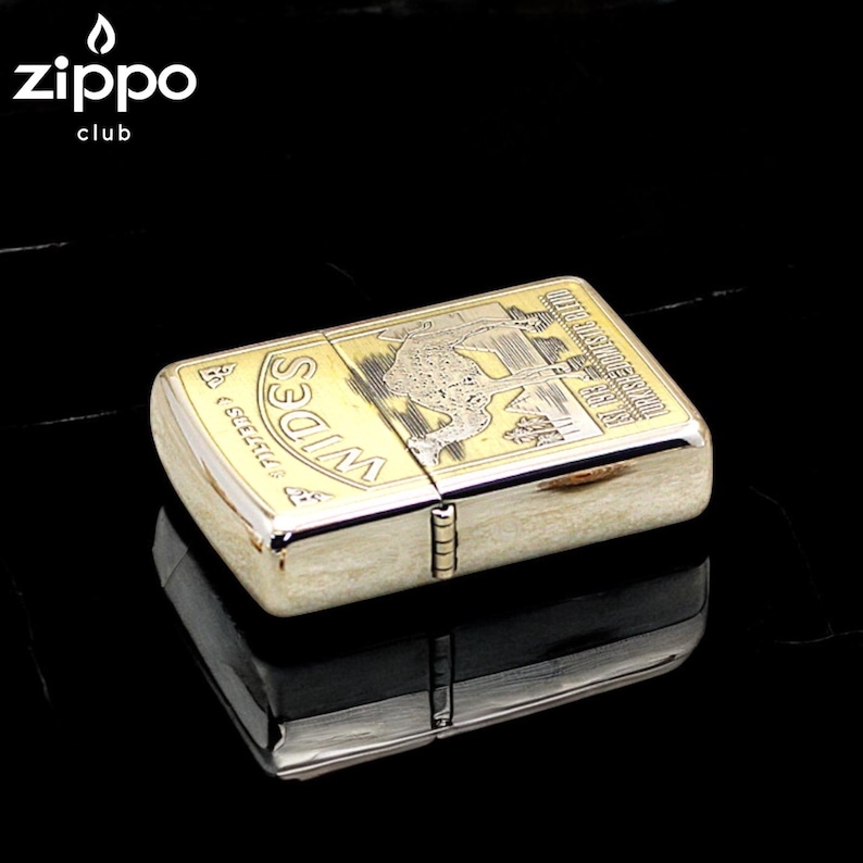 Pode incluir: Um isqueiro Zippo dourado com um design gravado detalhado. O isqueiro tem formato retangular com bordas arredondadas e uma tampa articulada. O design inclui uma imagem de um leopardo e o texto "WIDES" e "TOURIST DOMESTIC RATIO". O logotipo do clube Zippo est&aacute; no canto superior esquerdo.