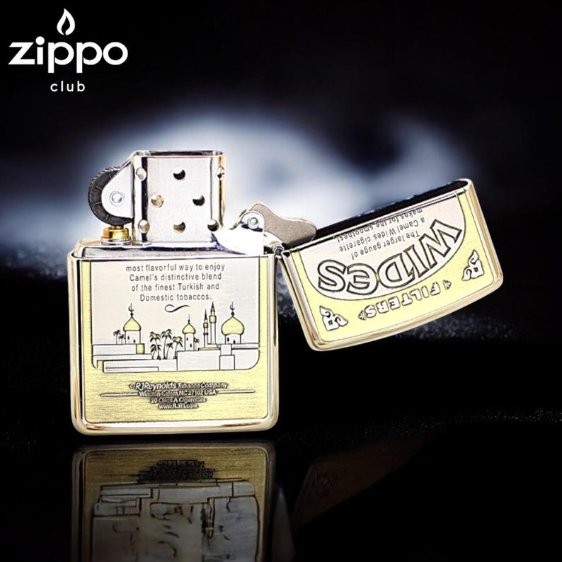 Pode incluir: Um isqueiro Zippo dourado, aberto para revelar o interior. O isqueiro apresenta texto e uma ilustra&ccedil;&atilde;o de edif&iacute;cios. A tampa exibe a palavra "Wides". O logotipo Zippo &eacute; vis&iacute;vel no canto superior esquerdo.