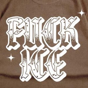 May include: Brown t-shirt with the white text "RUCK ICE" in a gothic font. Two white starbursts are on the top right and left of the text. The shirt is made of a soft material.