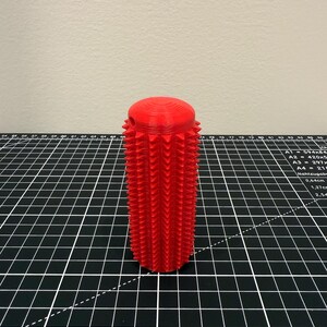May include: A bright red, cylindrical object with a textured surface. The object has a rounded top and is covered in rows of triangular spikes. The item is likely a decorative or functional piece, possibly a handle or knob.