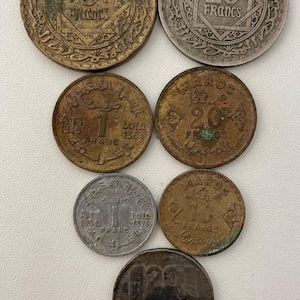 Puede incluir: Una colección de monedas marroquíes antiguas, con denominaciones como 5, 10 y 20 francos. Las monedas muestran diversos grados de desgaste, con texto visible y diseños intrincados. La colección incluye monedas de bronce y plata.