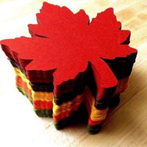 May include: A stack of colorful, maple leaf-shaped paper cutouts. The top leaf is a vibrant red, with layers of orange, yellow, green, and black visible below. These decorative paper leaves are perfect for fall-themed crafts and decorations.