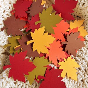 May include: A collection of colorful paper maple leaves in shades of red, orange, yellow, and brown. The leaves are scattered on a cream-colored, crocheted surface, creating a fall-themed decorative arrangement.