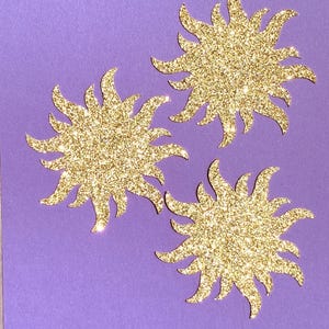 May include: Three gold glitter sunburst cutouts on a purple background. Each sunburst has multiple pointed rays extending outwards. The glitter catches the light, creating a sparkling effect. The sunbursts are arranged in a slightly offset pattern.