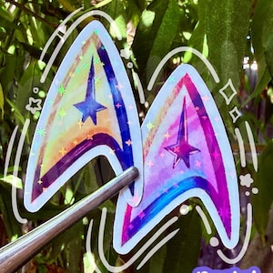 Puede incluir: Dos pegatinas holográficas con forma de insignias delta, una con tonos de acuarela azul y amarillo y la otra con tonos rosa y morado. Ambas pegatinas tienen una estrella central y un borde blanco con detalles de líneas blancas dibujadas a mano.