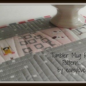 May include: A grey and pink quilted mug rug with a white mug on top. The rug has a geometric pattern and the text "Tumbler Mug Rug Pattern... by eamylove" is printed on the bottom right corner.