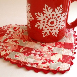 以下が含まれることがあります： 赤いハート型のキルトコースターで、縁はギザギザになっています。白い雪の結晶模様の赤いマグカップがコースターの上に置かれています。