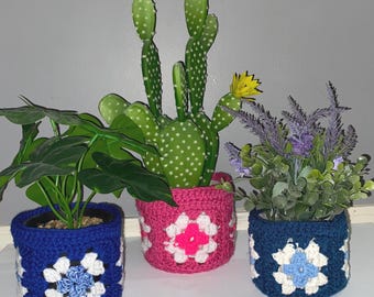 May include: Three crochet plant pot covers in blue, pink, and navy. Each pot has a white and colored floral design. The pots hold artificial plants, including a cactus with yellow flowers and a lavender plant.