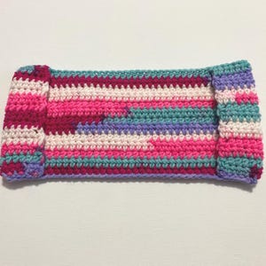 May include: A handmade crochet headband in a rectangular shape. The headband features a colorful striped pattern with shades of pink, purple, teal, and white. The ends are folded over, creating a finished look.
