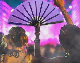 May include: A purple hand fan with black accents is held up in the air. The fan has a glittery border and is open, displaying its full shape. The background is a blurred scene with bright lights, suggesting a concert or festival setting.