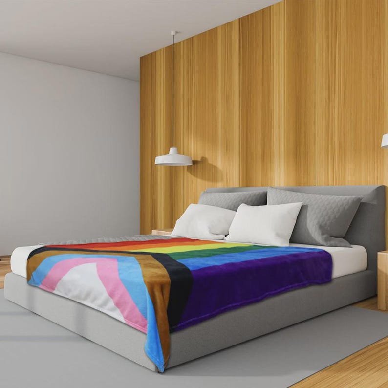 K&ouml;nnte beinhalten: Eine weiche Decke mit dem Progress Pride Flaggen-Design, mit Regenbogen-, braunen, schwarzen, rosa, hellblauen und wei&szlig;en Streifen. Die Decke liegt auf einem Bett mit wei&szlig;en Kissen und einem grauen Kopfteil in einem modernen Schlafzimmer.