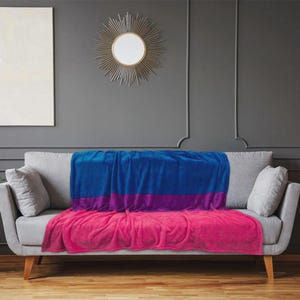 K&ouml;nnte beinhalten: Eine weiche Decke in Blau-, Lila- und Rosat&ouml;nen liegt auf einem grauen Sofa. Die Decke ist so gefaltet, dass die Farben zur Geltung kommen, mit Blau oben, einem lila Streifen in der Mitte und Rosa unten. Zwei graue Kissen liegen auf dem Sofa.