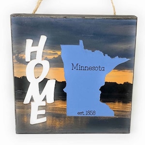 Peut inclure: Une pancarte en bois avec un fond de coucher de soleil et une silhouette blanche de l'état du Minnesota. La pancarte indique "Home" avec le nom de l'état et "est. 1858" en dessous.