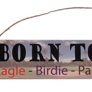 May include: A rectangular wooden sign with the text "BORN TO GOLF" in large black letters. Below, the words "Eagle - Birdie - Par - Bogey - Beer" are written in different colors. The sign has a golf course background and a twine hanger.