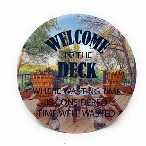 May include: Round metal sign with a scenic deck image, featuring Adirondack chairs and the text "WELCOME TO THE DECK WHERE WASTING TIME IS CONSIDERED TIME WELL WASTED." Includes a hole for hanging.