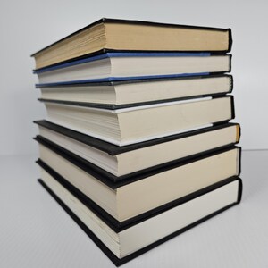 May include: A stack of seven hardcover books with black covers and off-white pages. The top book has tan pages, and the second book has a blue spine. The books are arranged in a neat pile on a white surface.