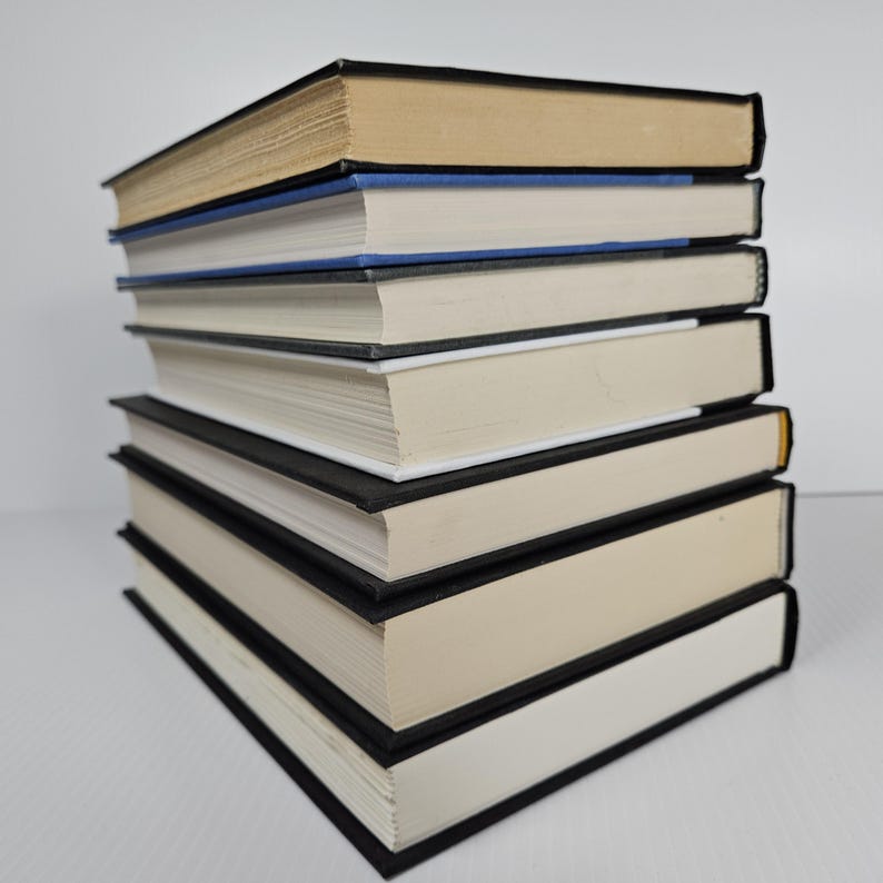 May include: A stack of seven hardcover books with black covers and off-white pages. The top book has tan pages, and the second book has a blue spine. The books are arranged in a neat pile on a white surface.