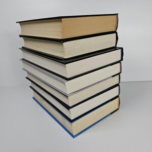 May include: A stack of hardcover books with black spines and varying colors. The top book has a tan cover, while the others have white or blue covers. The pages are cream-colored, and the books are neatly stacked.