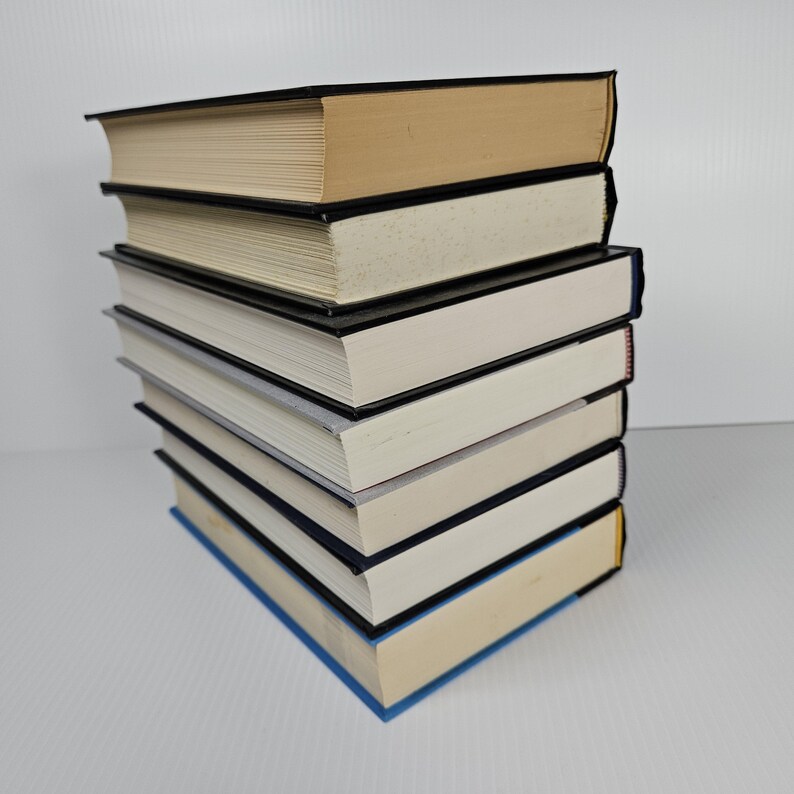 May include: A stack of hardcover books with black spines and varying colors. The top book has a tan cover, while the others have white or blue covers. The pages are cream-colored, and the books are neatly stacked.