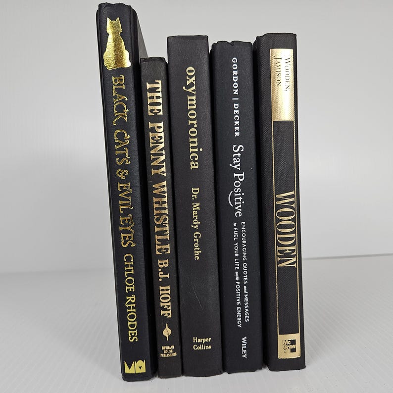 May include: A collection of five black books with gold lettering. Titles include "Black Cats & Evil Eyes," "The Penny Whistle," "Oxymoronica," "Stay Positive," and "Wooden." The books are arranged vertically, with the spines facing the viewer.