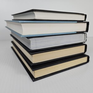May include: A stack of five hardcover books with black covers and white or cream-colored pages. The top book has a black cover, and the second book has a light blue cover. The books are stacked on a white surface.